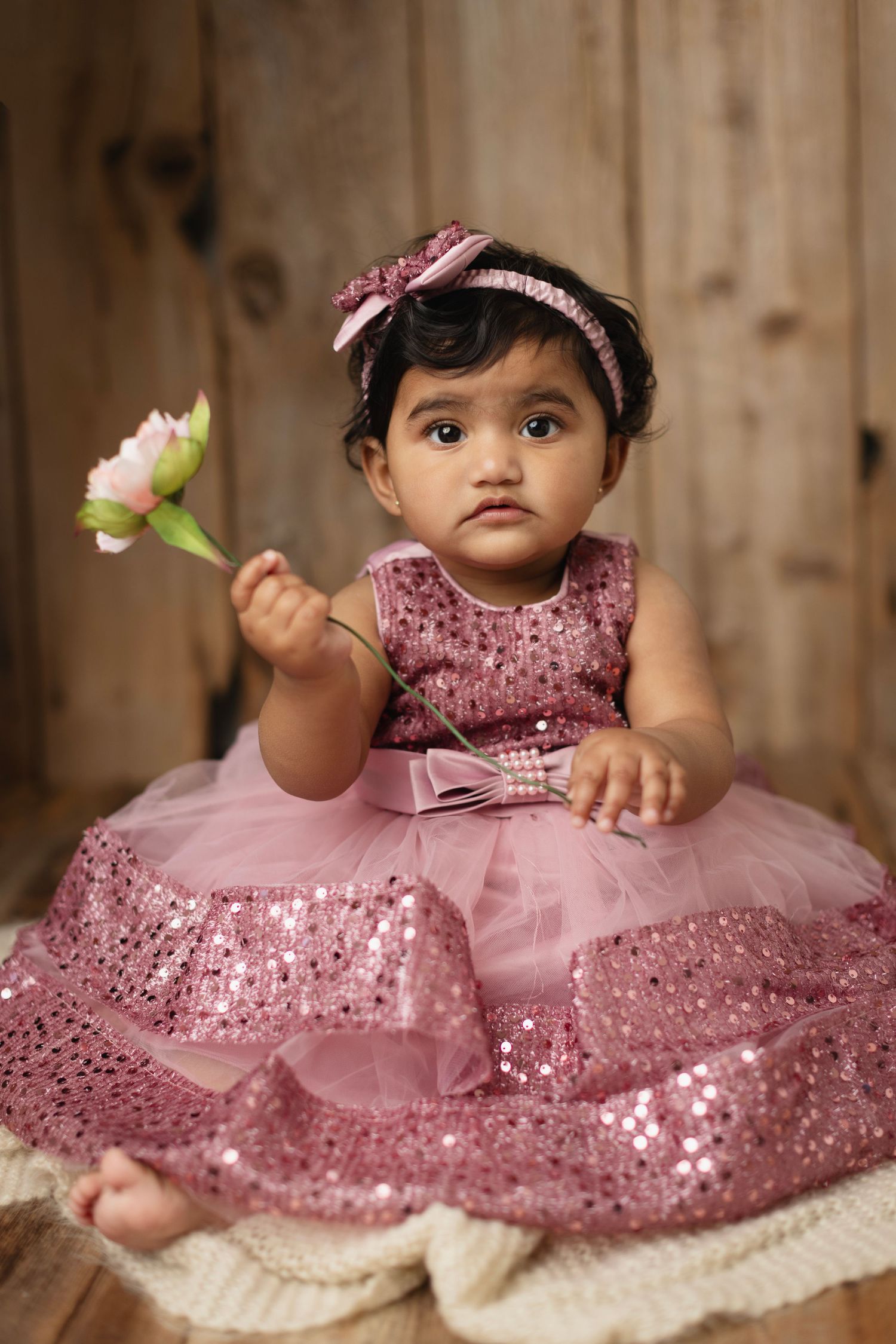 Baby girl in pink sequin tutu dress with matching headband holds a flower, sitting on wooden floor.