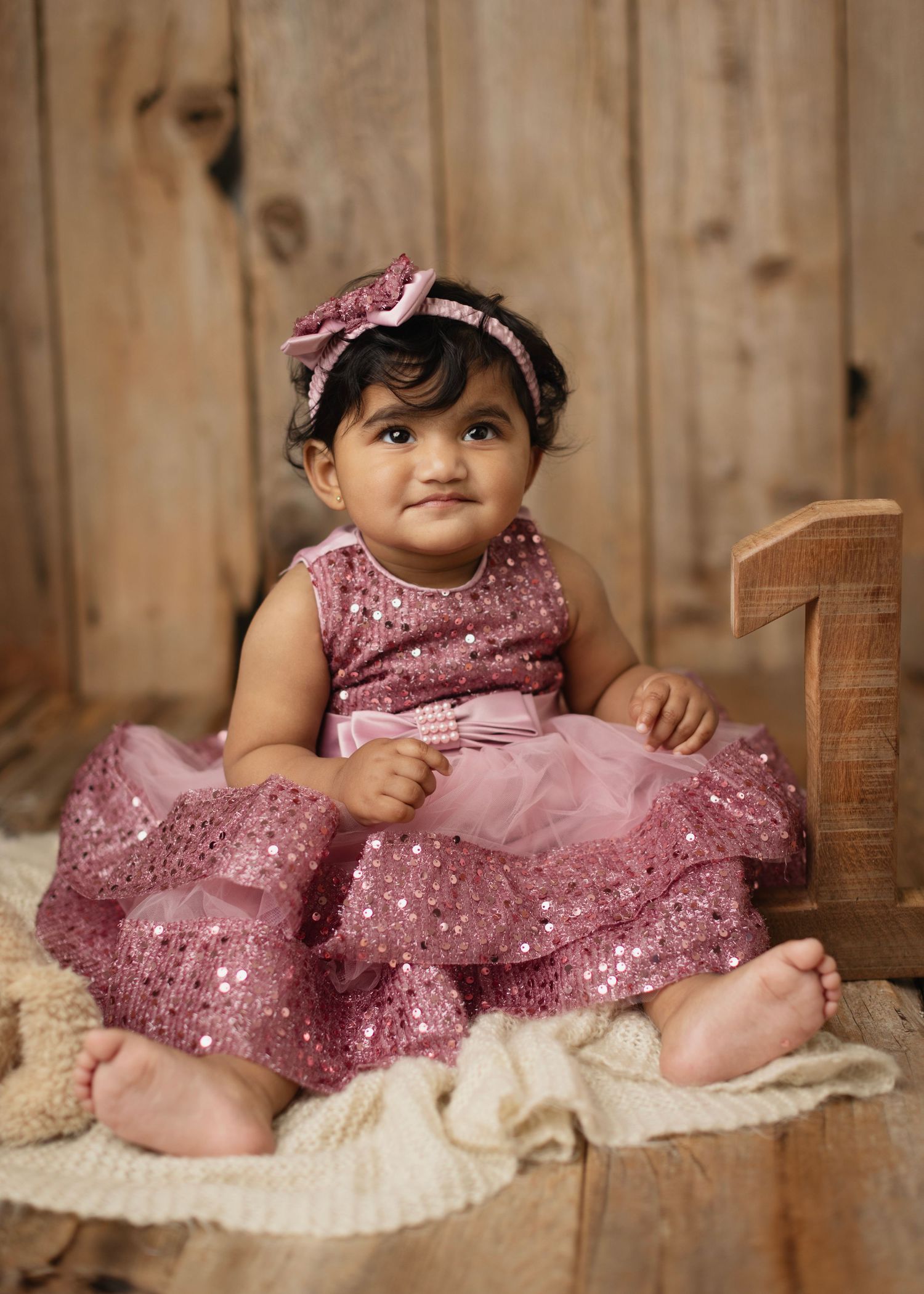 Adorable baby girl in a sparkly pink dress and bow headband sitting beside a wooden number one.