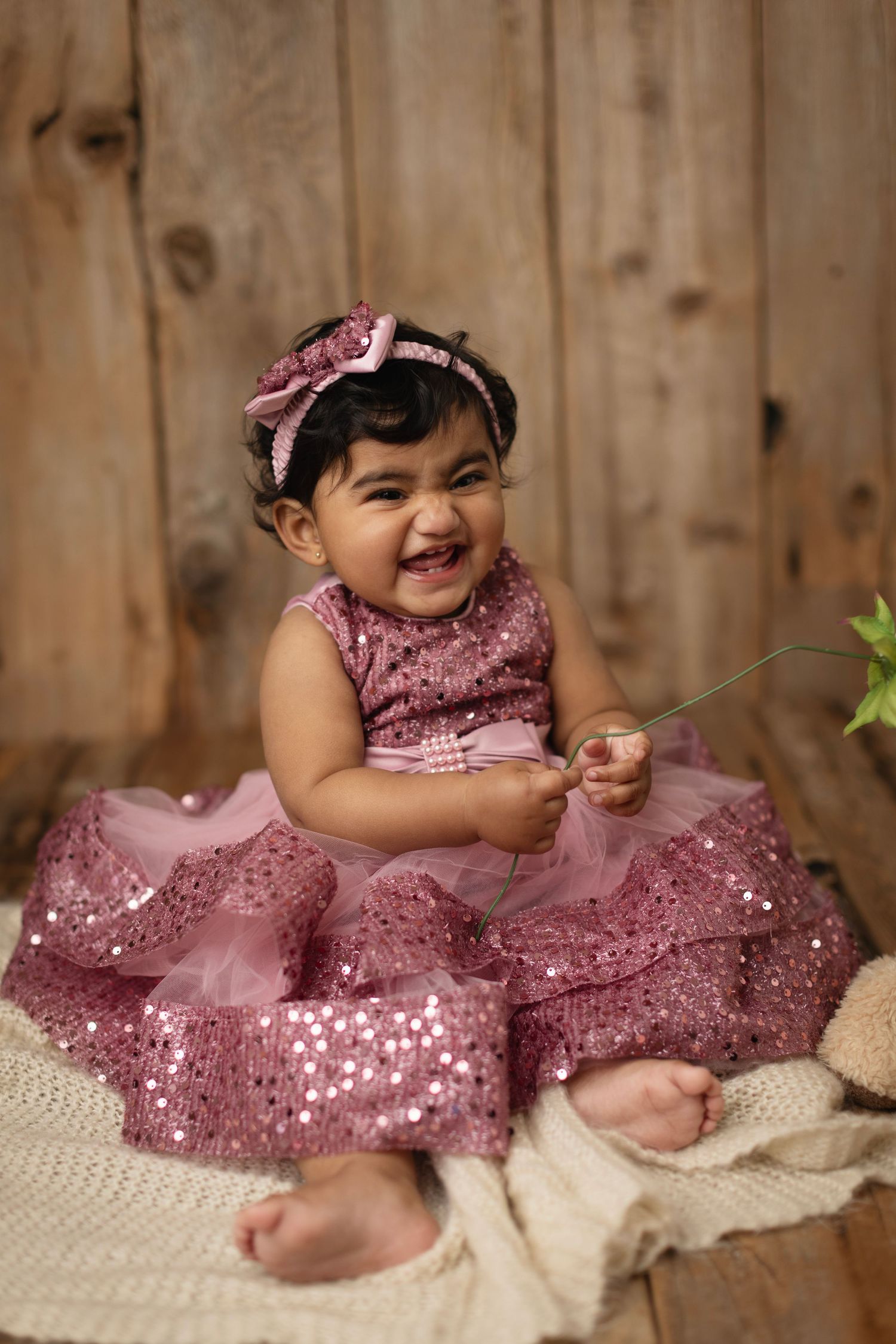 A joyful baby girl in a sparkly pink sequin dress and matching headband laughs while sitting on a blanket.