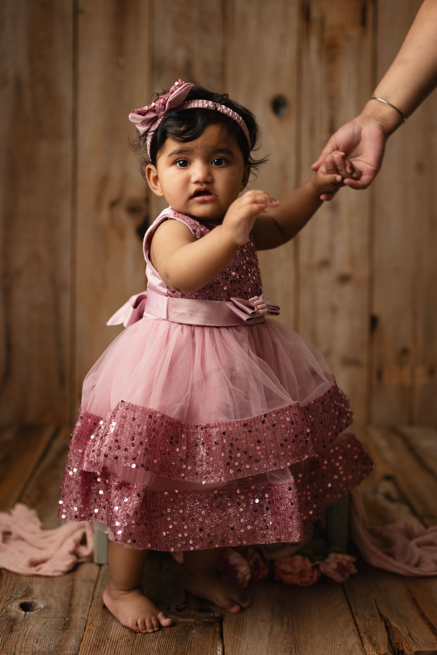 A baby girl in a pink sequined tiered dress with bow and matching headband stands holding an adult's hand on a wooden backdrop.