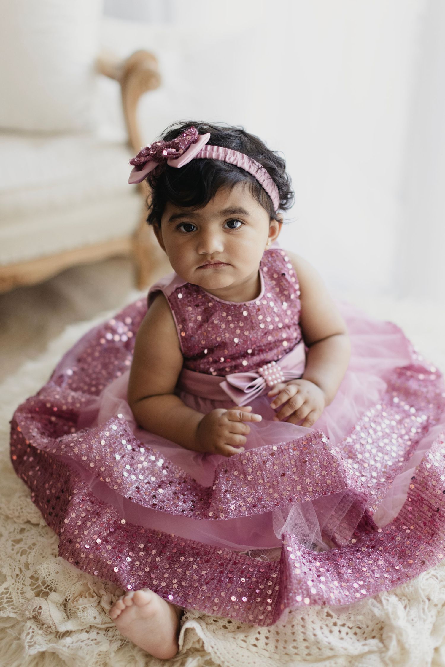 Baby girl in pink sequin tulle dress with matching bow headband sits on cream blanket.