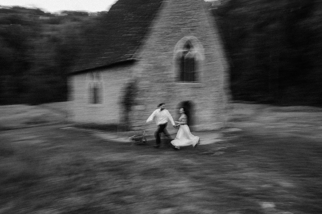 Blurred black and white photo of couple running hand in hand past a stone chapel.