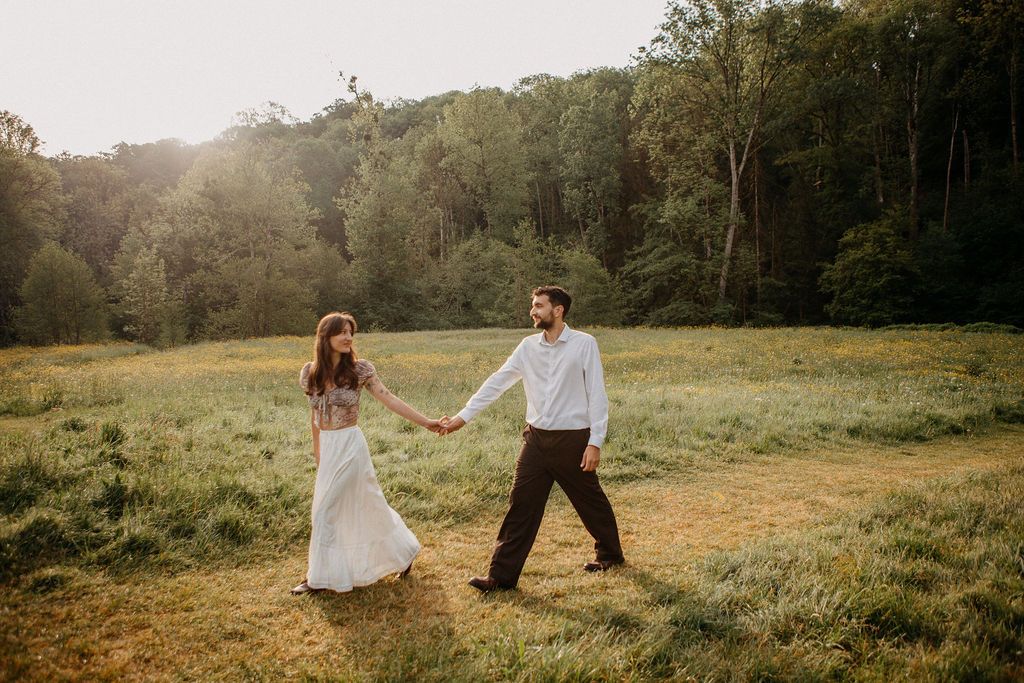 Couple holding hands walking through a sunlit green meadow surrounded by lush forest.