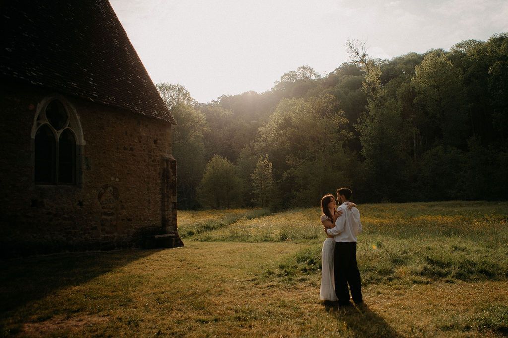 Couple embracing near a stone chapel at golden hour with forest backdrop at sunset.