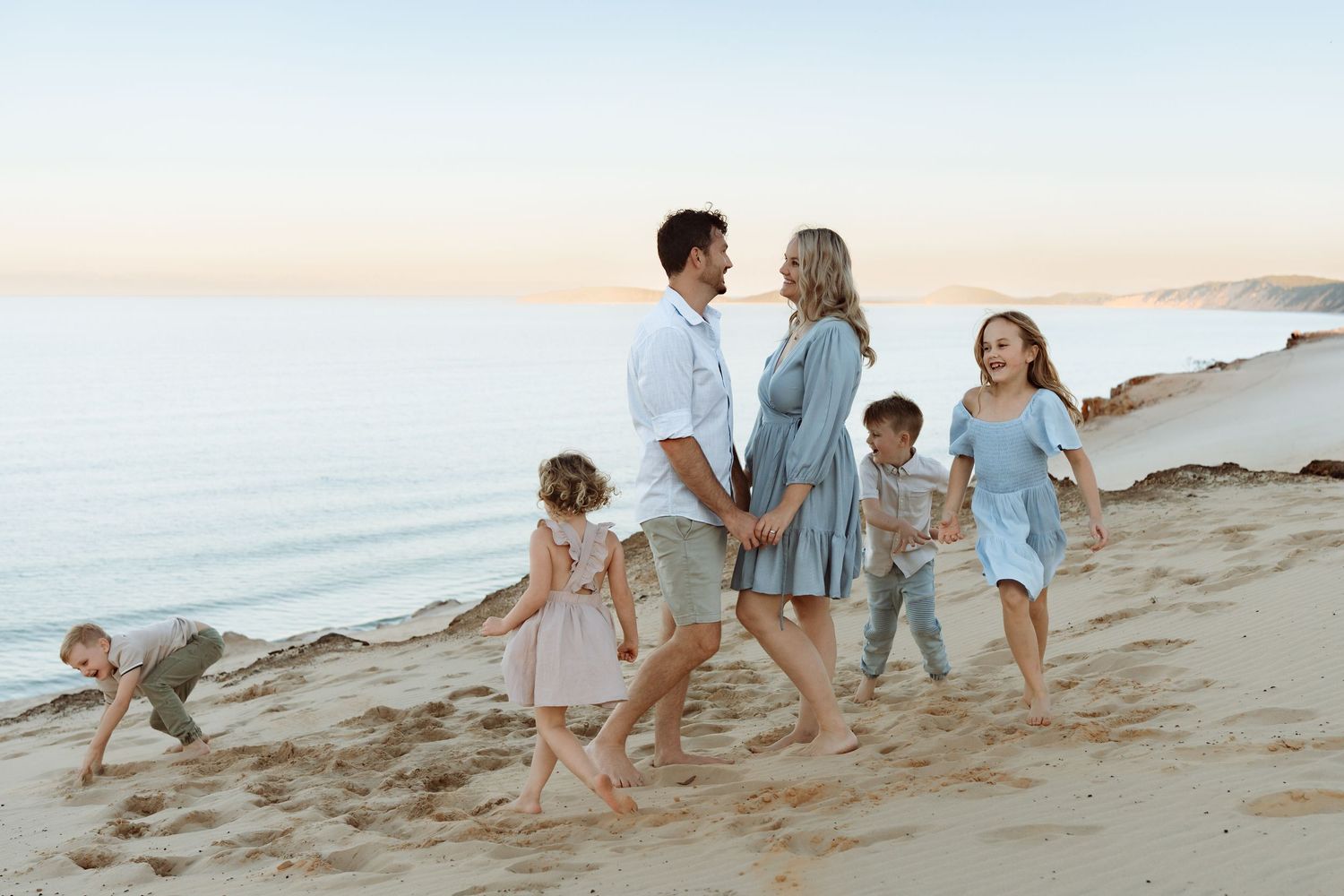 Family walking on sandy beach at sunset, parents holding hands while children play around them near calm ocean waters. Carlo Sandblow