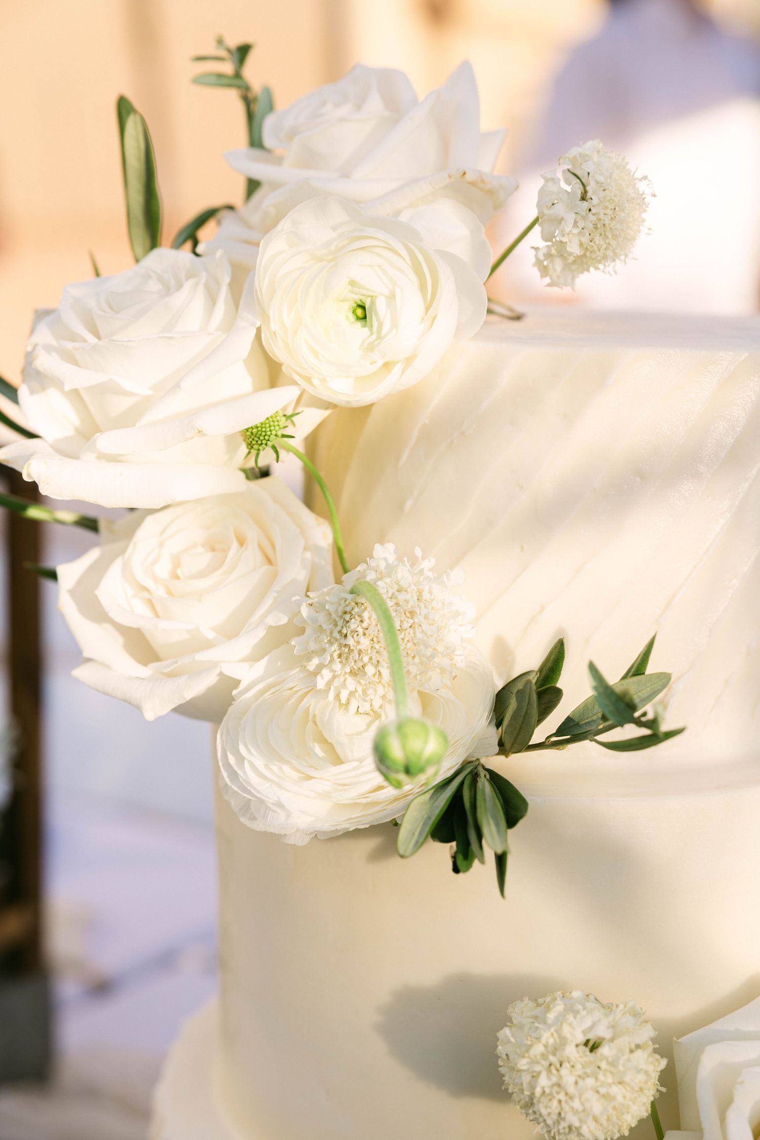Elegant white wedding cake adorned with fresh white roses, ranunculus, and green foliage floral decorations.