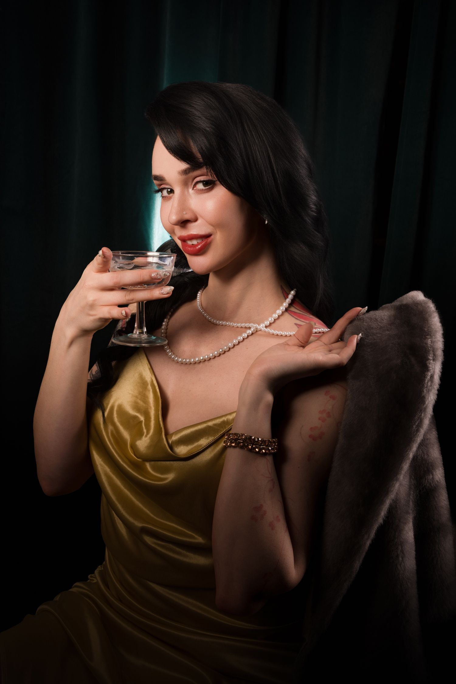Elegant woman in gold dress and pearl necklace holding a cocktail glass, draped in fur coat against dark green backdrop.