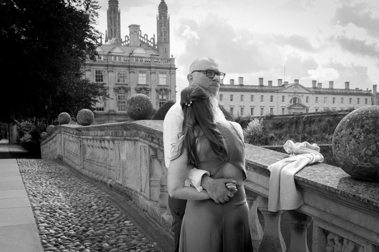 Black and white photo of a couple embracing on a stone bridge near a grand historic building.