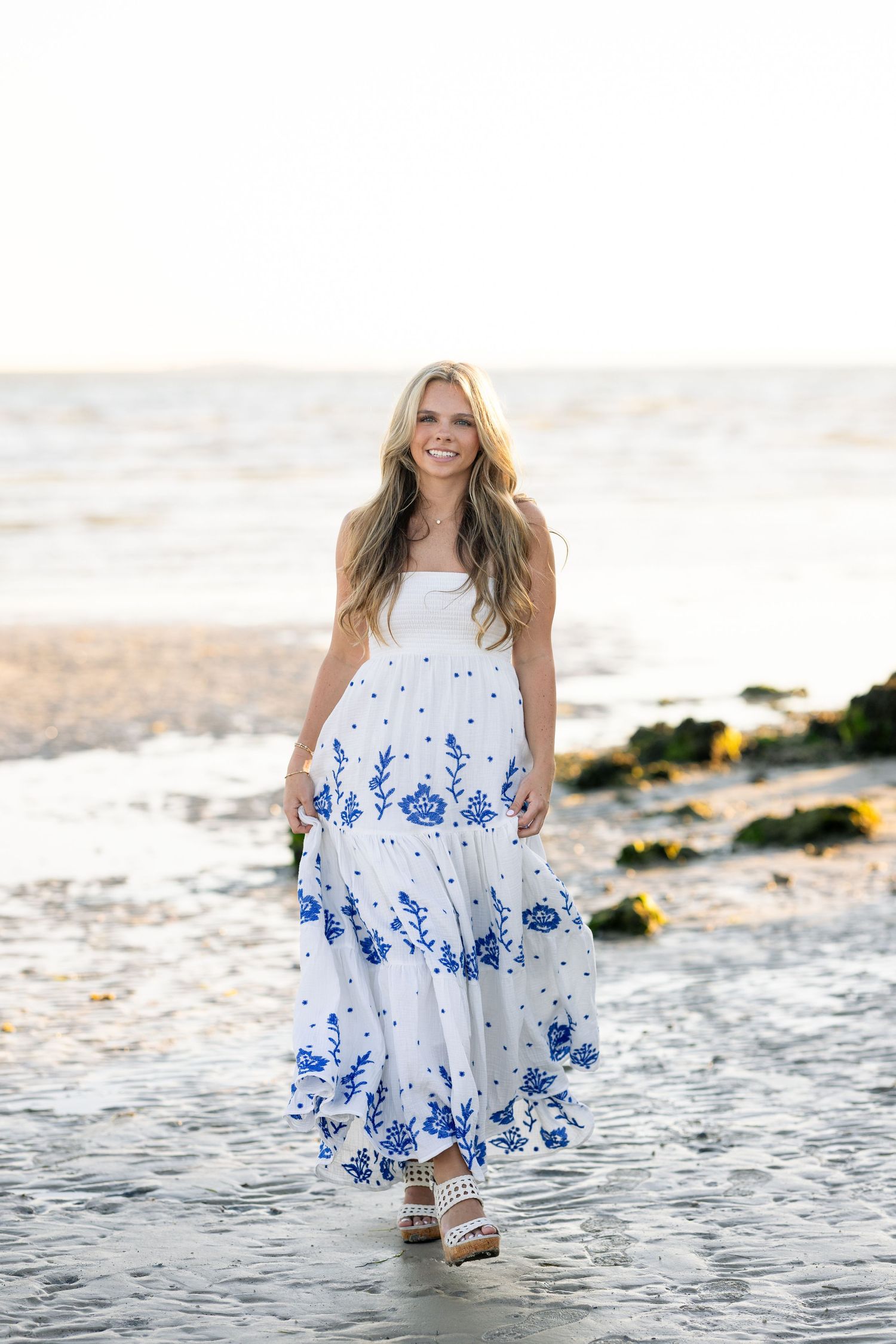 Smiling blonde woman in white and blue floral maxi dress walking barefoot on a sandy beach at sunset.