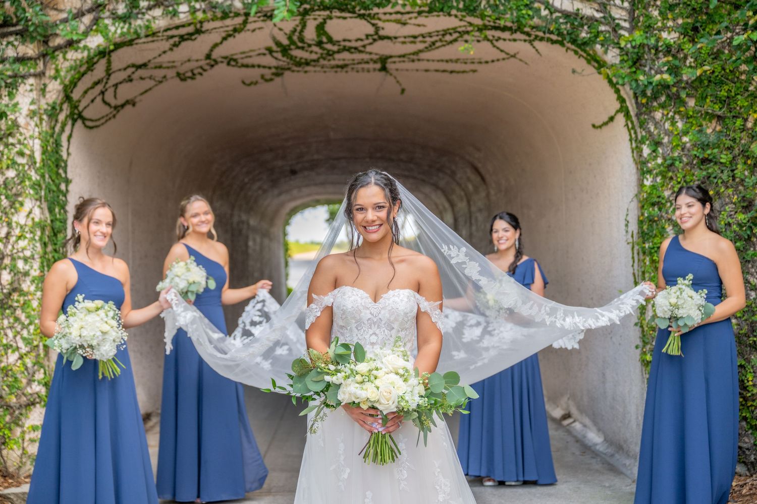 Bride in white gown holds veil wide flanked by four bridesmaids in blue dresses under ivy-covered stone archway.