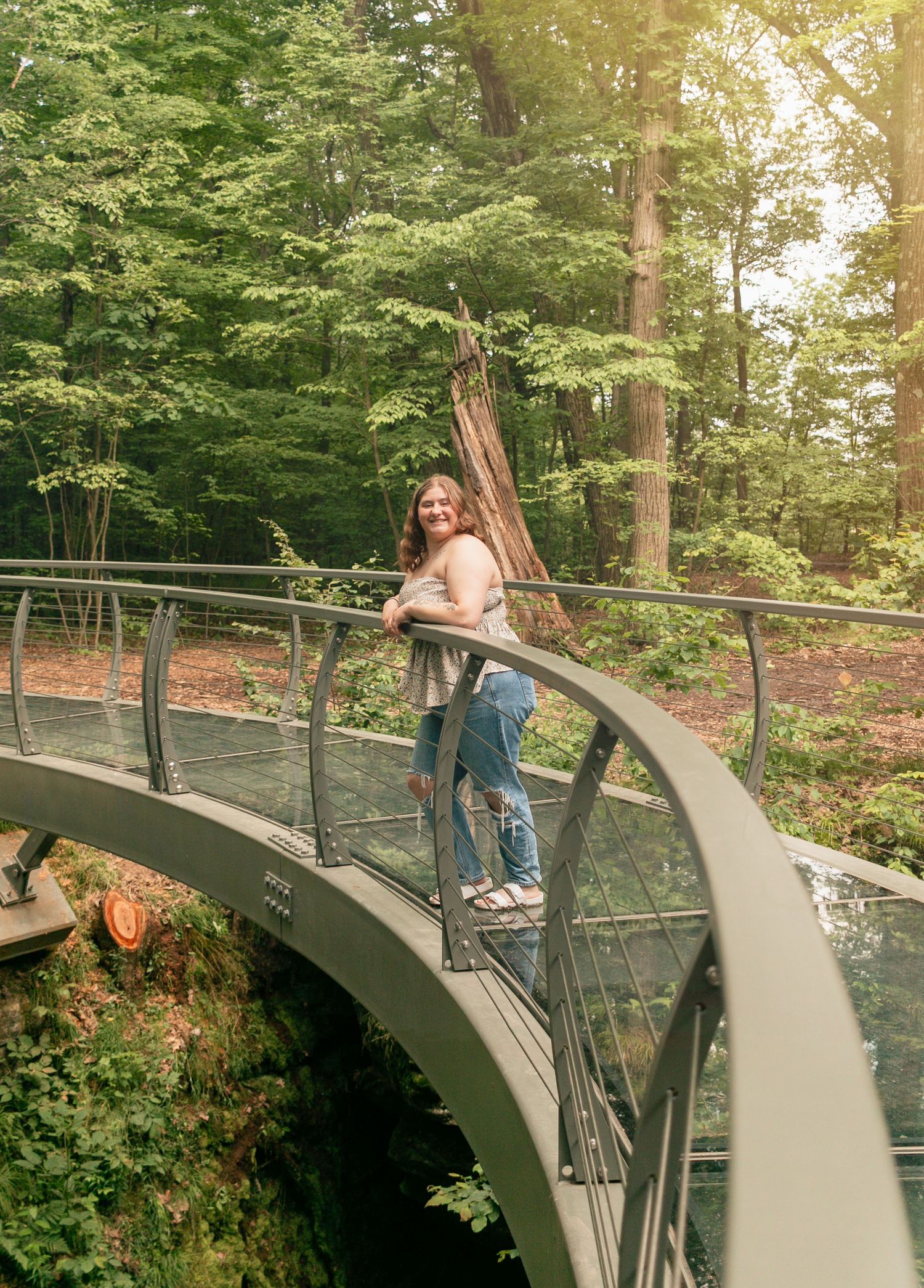 Woman in jeans posing on a curved metal bridge surrounded by lush green forest trees.