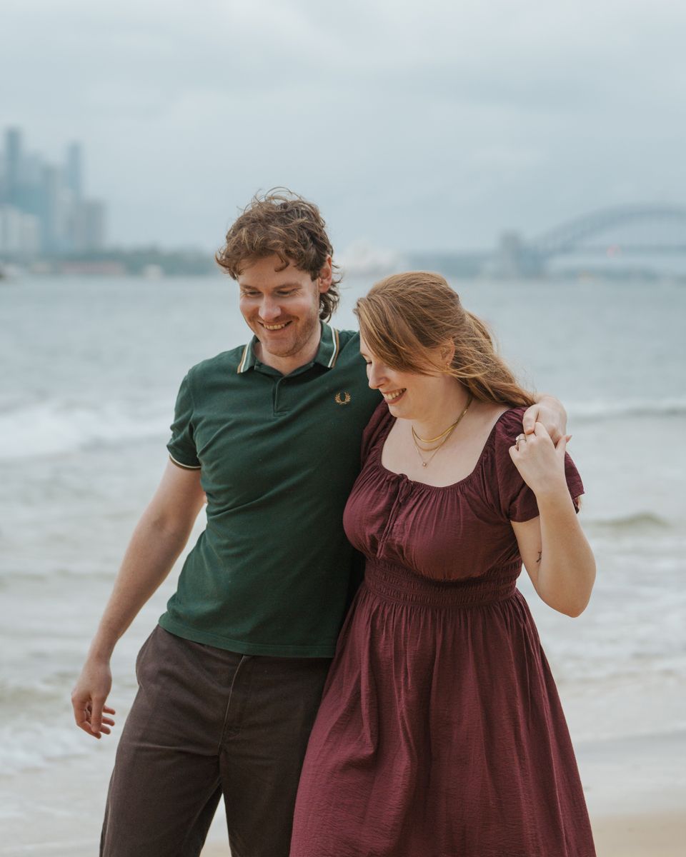 Couple smiling and walking on a beach with Sydney Harbour Bridge in the background on a cloudy day during an engagement photoshoot