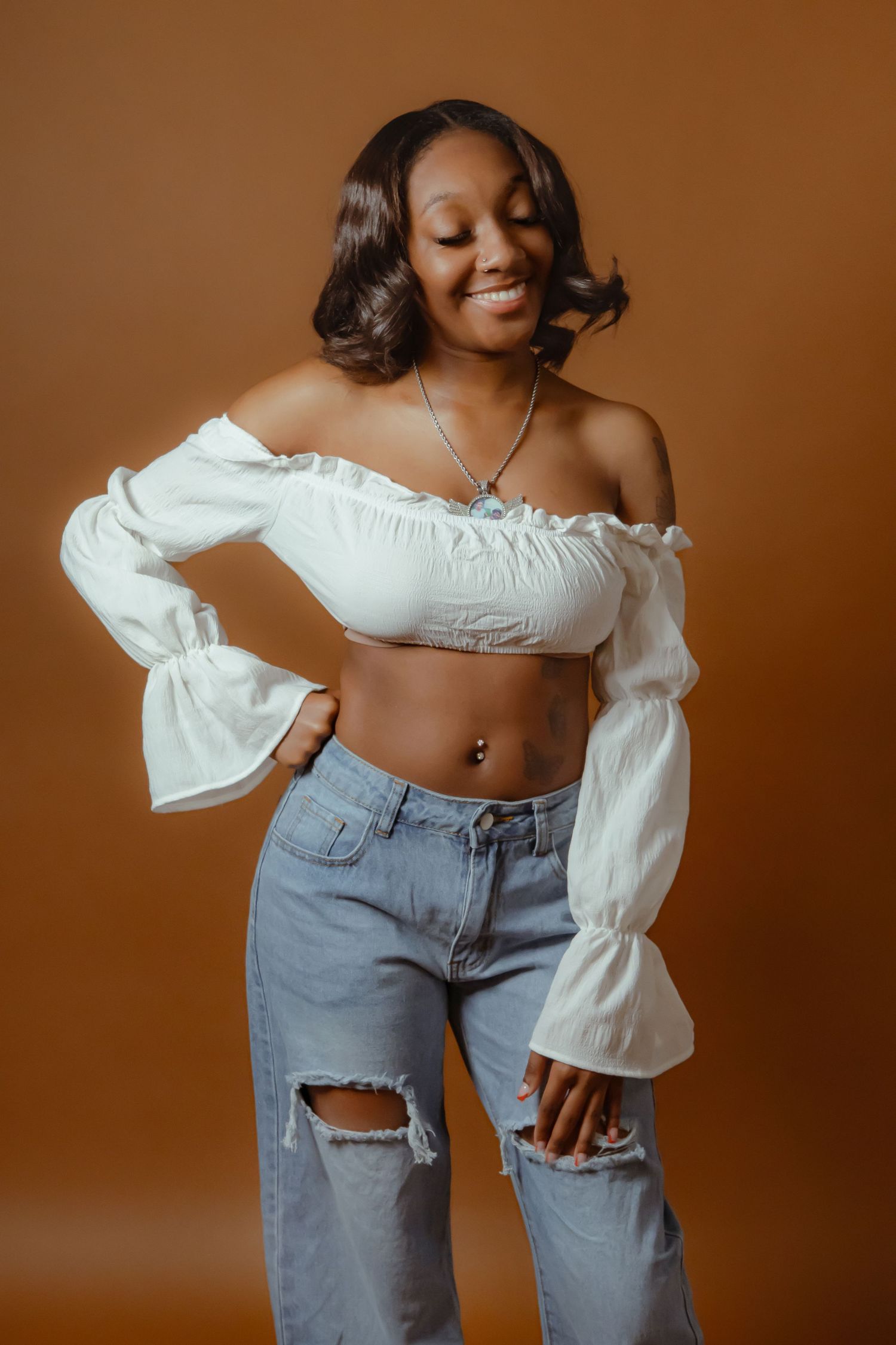 Smiling young woman in white off-shoulder crop top and ripped jeans posing against a warm brown background.