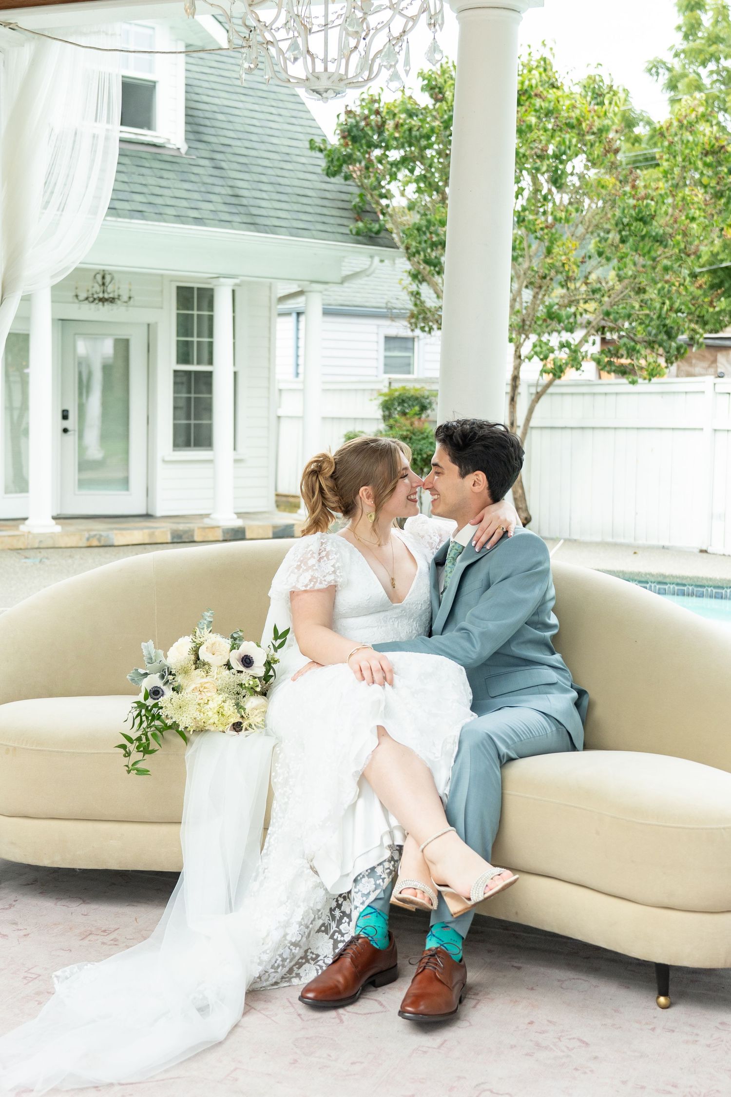 Bride and groom sit closely on a cream sofa, sharing a tender moment, with a white bouquet and elegant venue backdrop.