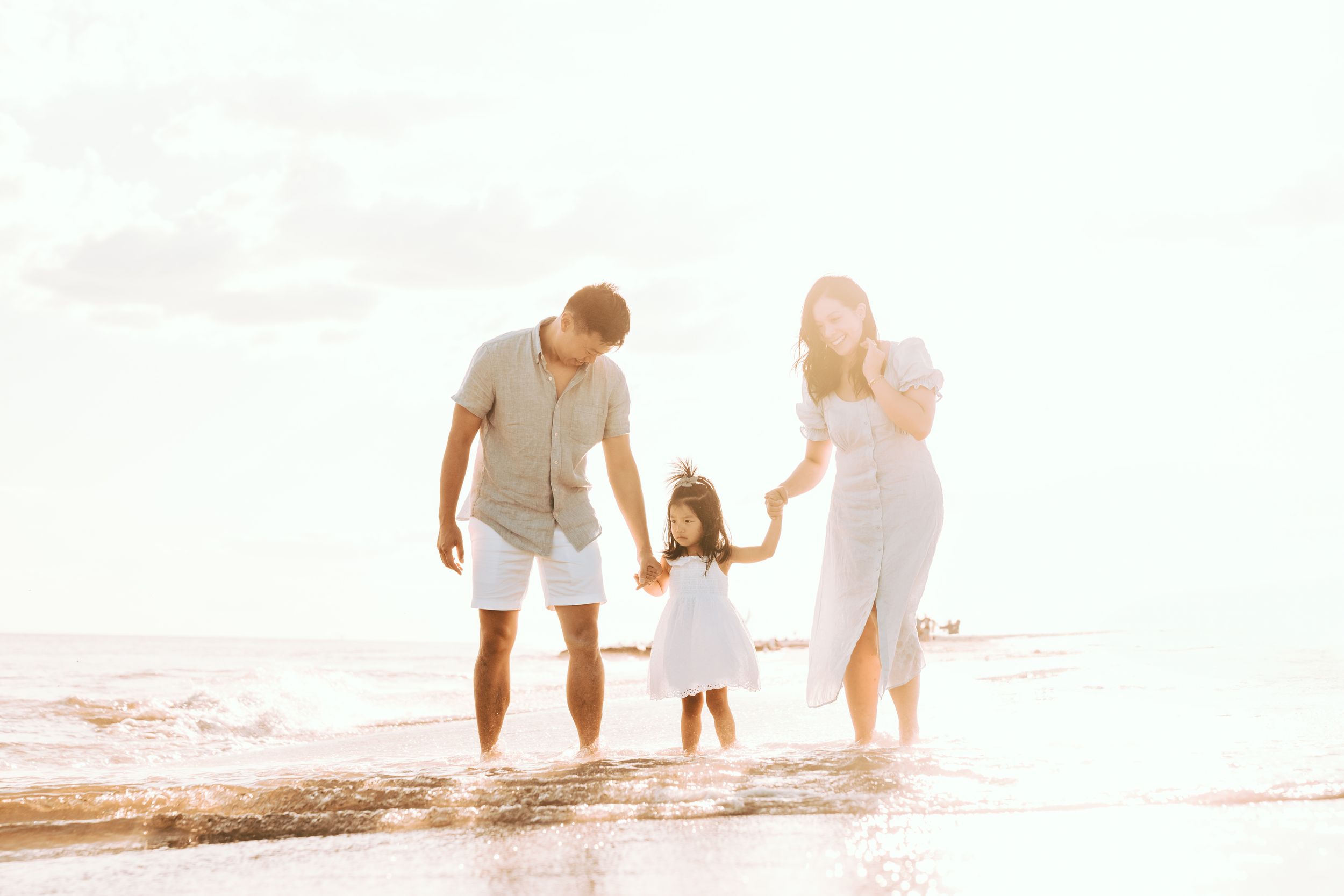 Happy family with young daughter walking on sunny beach shore of Long Beach, waves washing over feet in golden light.