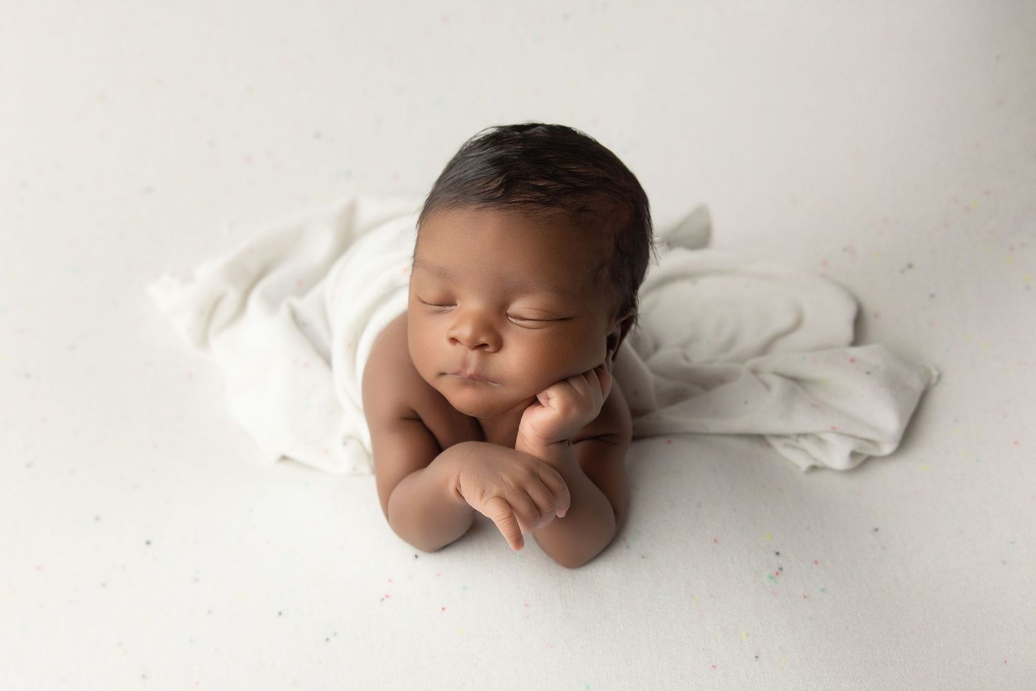 A peaceful newborn baby sleeping on a white backdrop, wrapped in a soft white cloth in a professional photo.