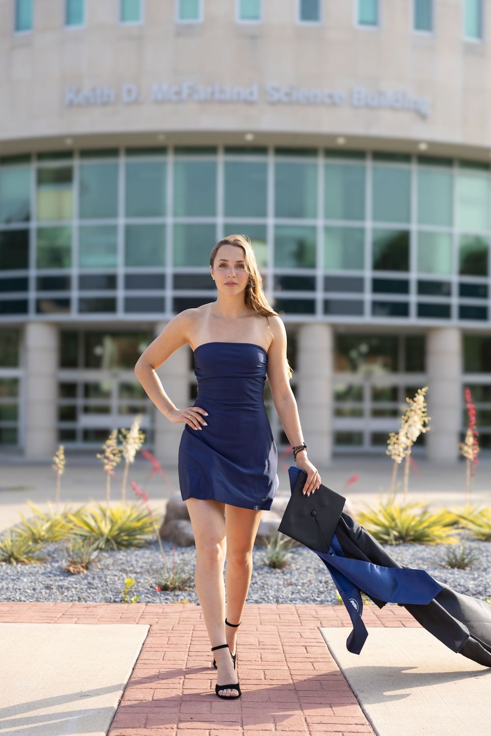 college grad walking away from the science building for the last time holding her graduation cap and gown