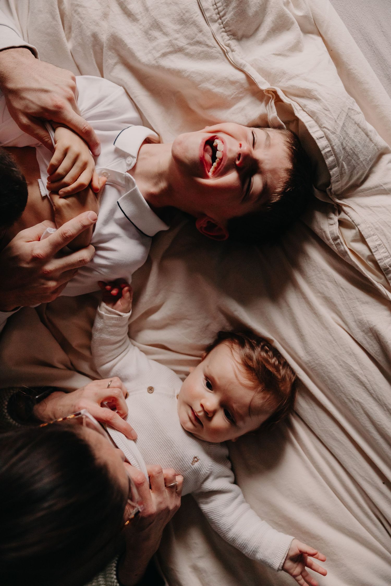 Overhead view of a family with two young children laughing and playing together on a cozy bed.