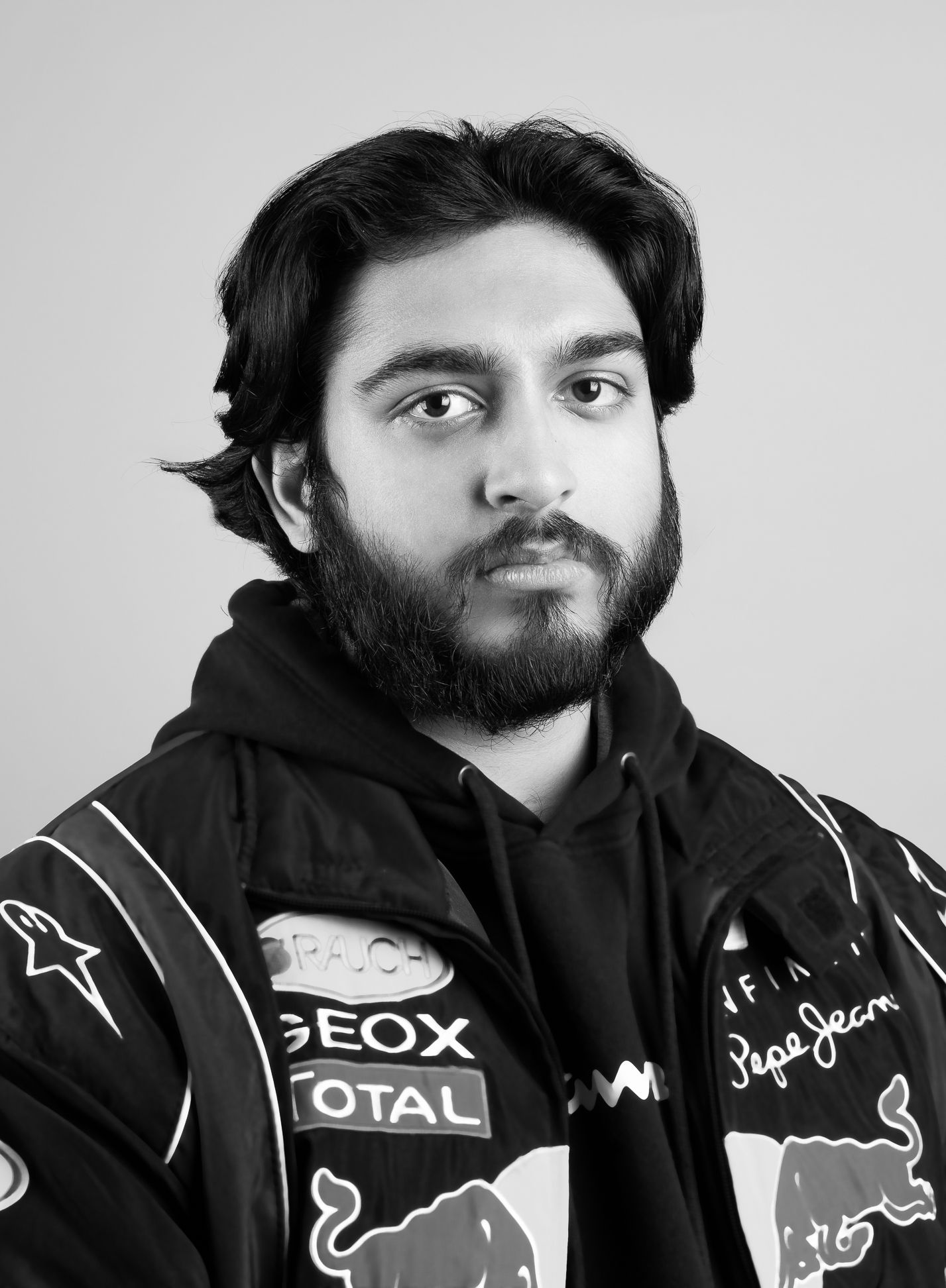 Black and white image of a man in a racing jacket and hoodie, with no smile, looking at the camera