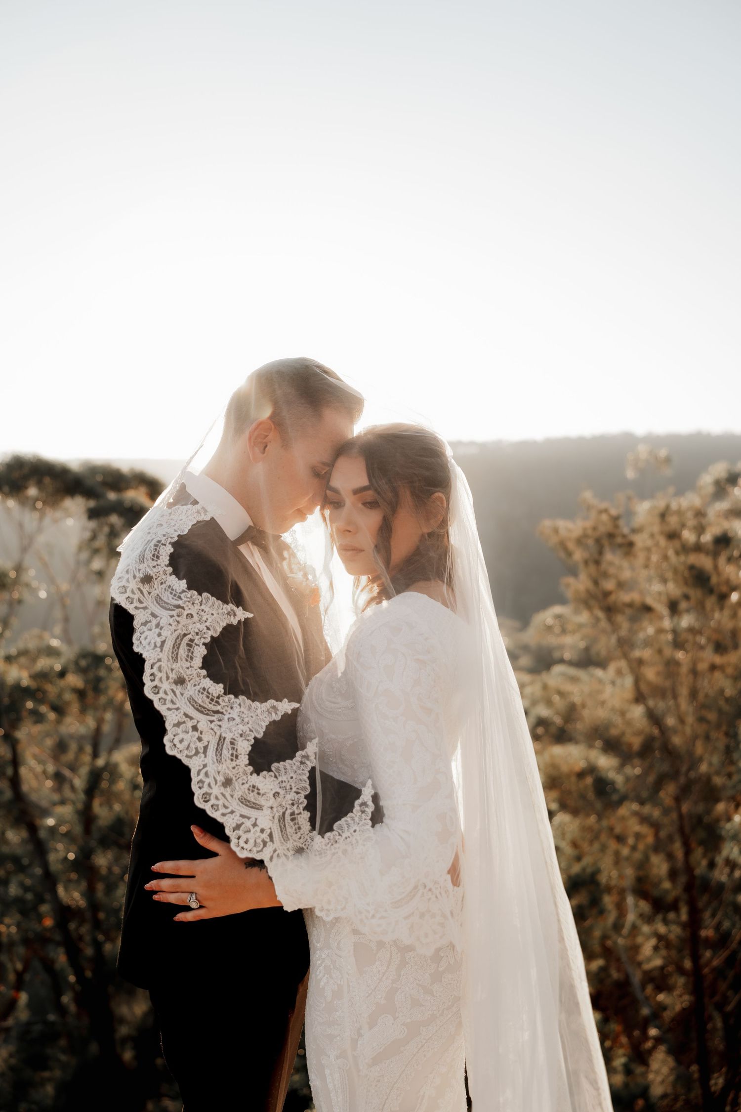 Bride and groom embrace outdoors at golden hour, bride wearing lace wedding dress and veil with scenic hills behind.
