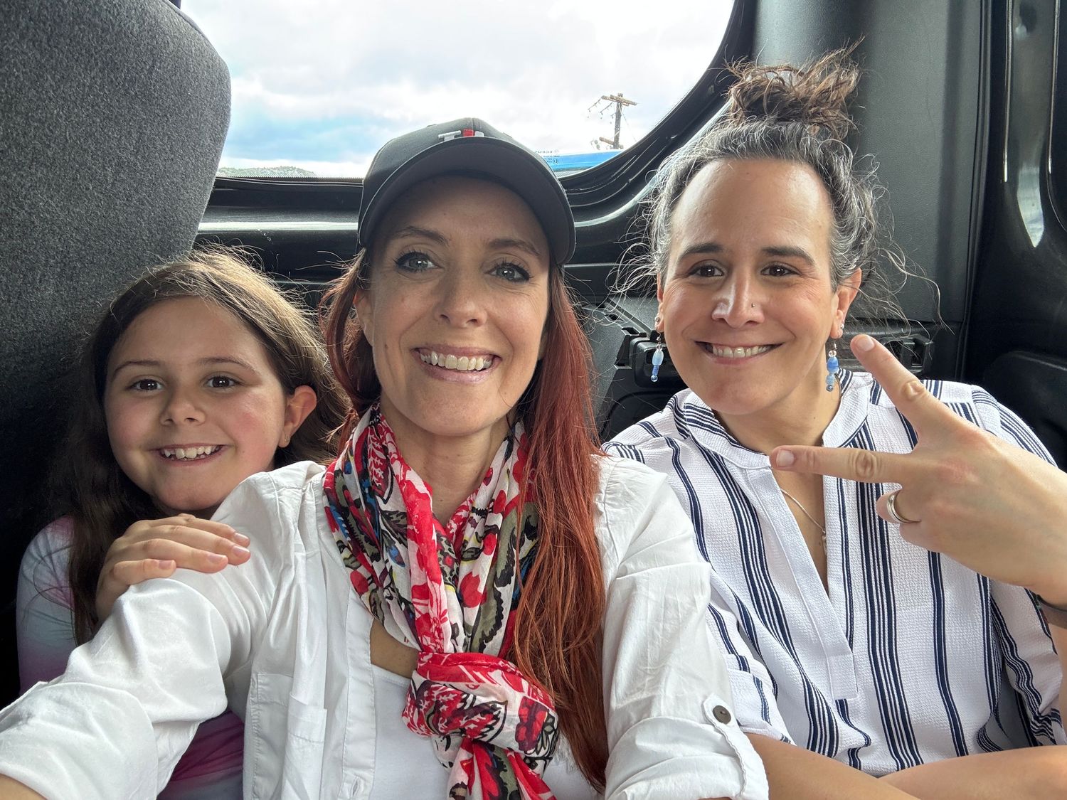 Three smiling women pose for a selfie inside a vehicle, with one wearing a cap and red scarf.