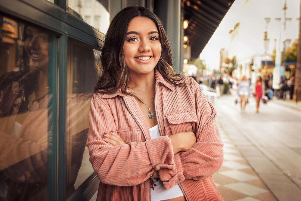 Young woman in pink jacket smiling with arms crossed on a sunny urban street.