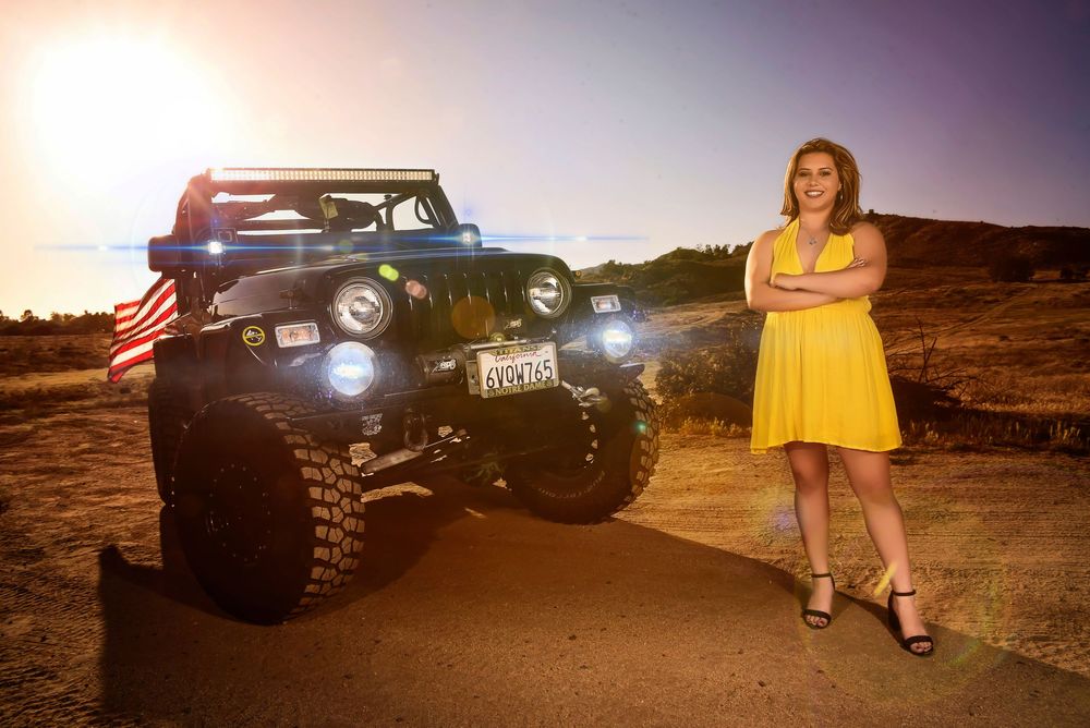 Young woman in yellow dress poses beside a lifted Jeep with American flag at sunset on a scenic overlook.