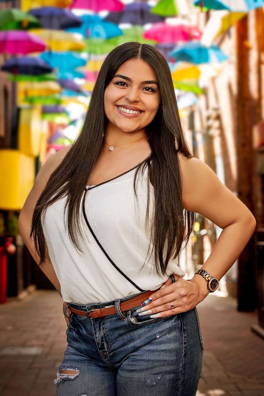 Smiling young woman in white top and jeans poses in colorful umbrella-lined alley.