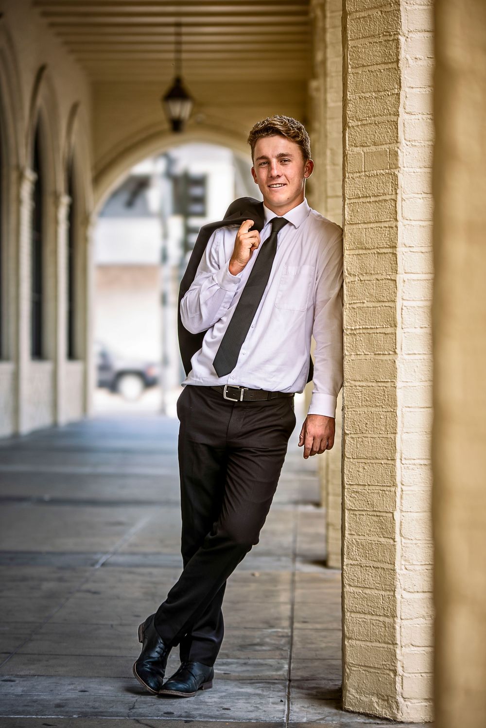 Young man in white shirt and tie leaning against brick column in arched corridor, holding jacket over shoulder.