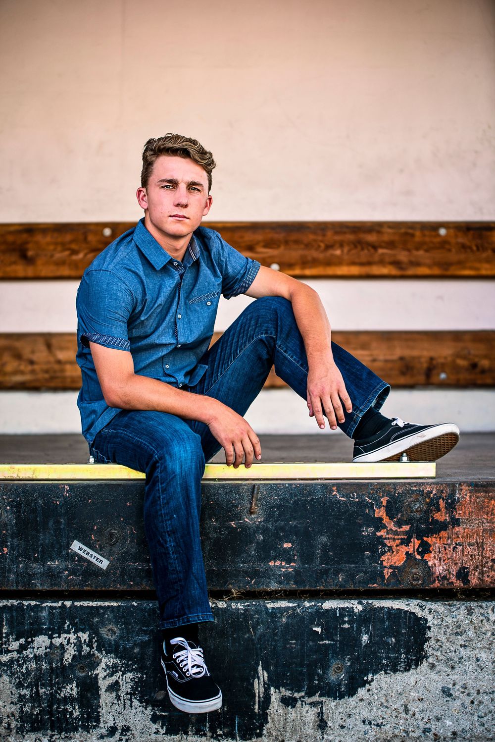 Young man in blue shirt and jeans sitting casually on bleachers wearing black Vans sneakers.
