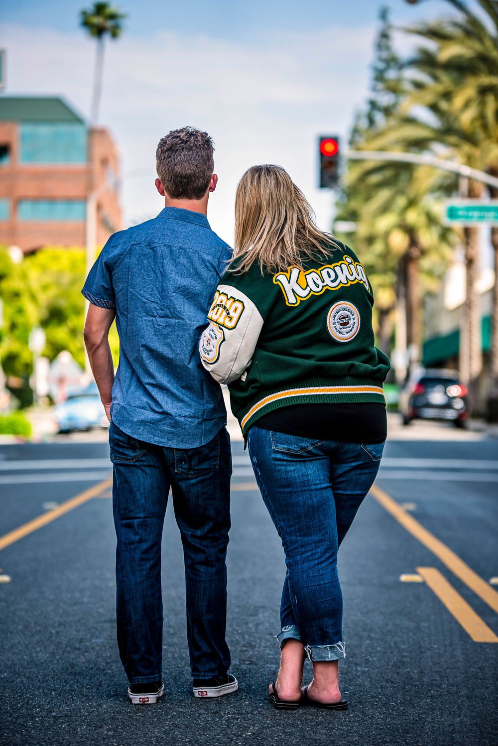 Couple standing in street, woman wearing green Koetter jersey holding football, man in blue shirt and jeans, palm trees background.