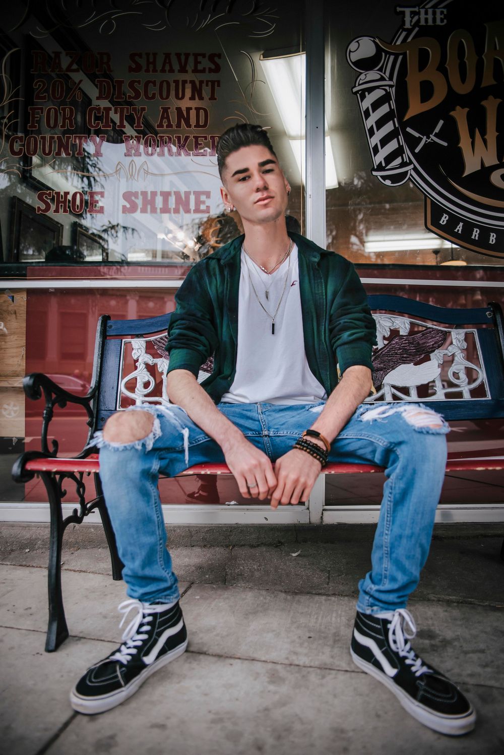 Young man in ripped jeans, green jacket, and high-top Vans sits on a bench outside a barbershop.