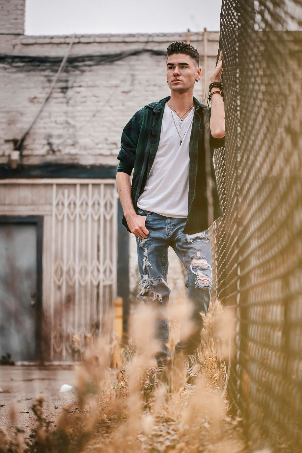 Young man in ripped jeans and green flannel shirt leaning against chain-link fence in urban setting.