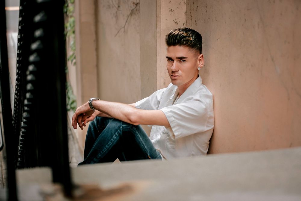 Young man in white shirt and jeans sitting relaxed against a beige wall, looking confidently at the camera.