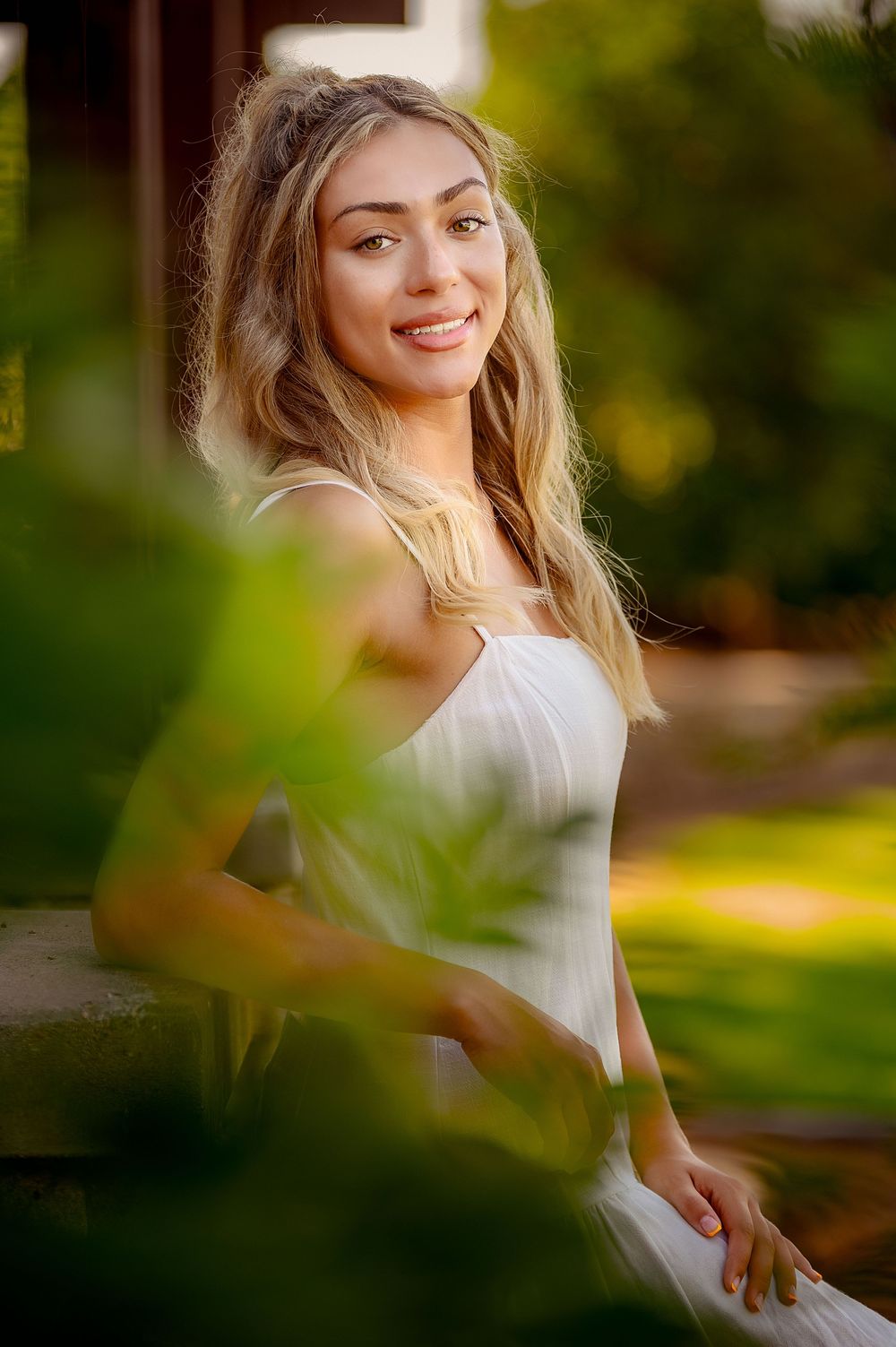 Young blonde woman in white dress smiling outdoors, surrounded by lush green foliage in warm sunlight.