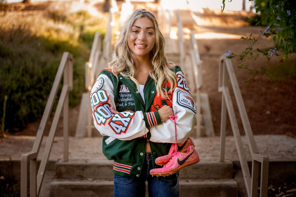 Young woman smiling in a varsity jacket holding pink sneakers outdoors on steps.
