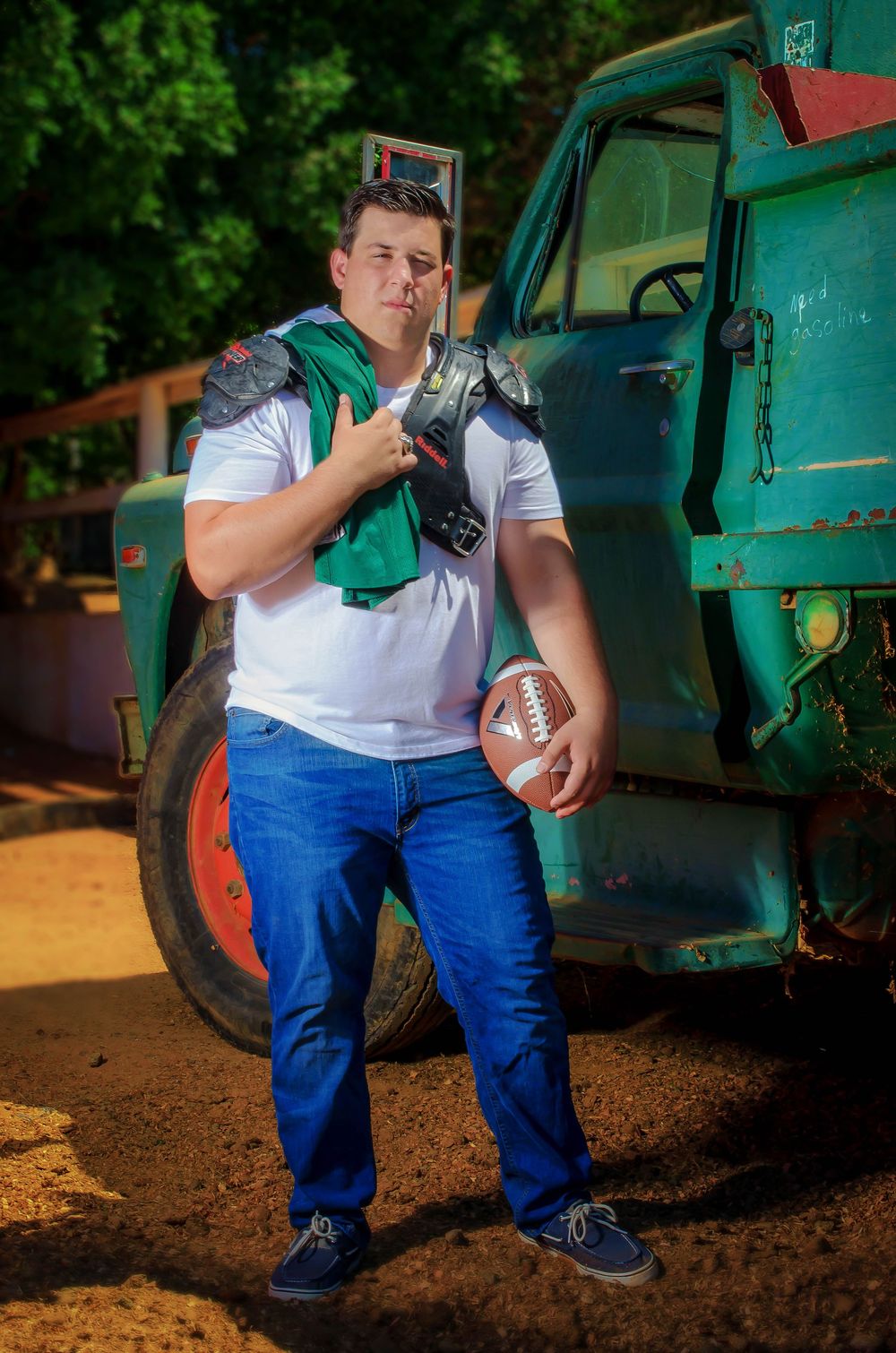 Young man in white shirt and jeans holds football wearing shoulder pads, leaning against vintage green truck outdoors.