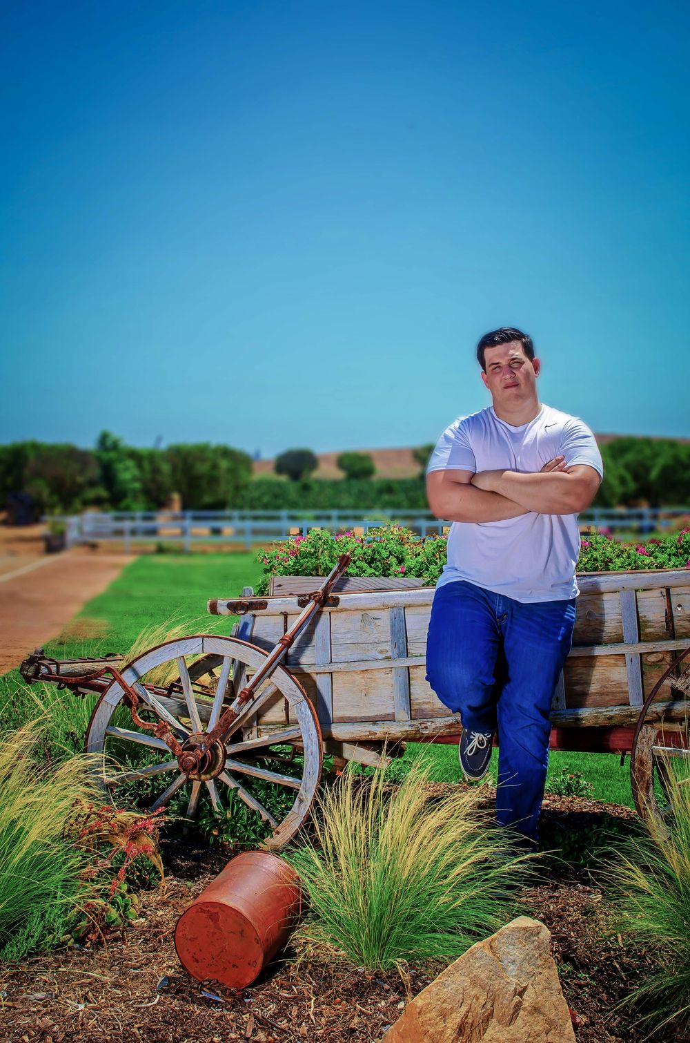 Man in white t-shirt and blue jeans leaning on a rustic wooden wagon with flowers in a scenic farm setting.