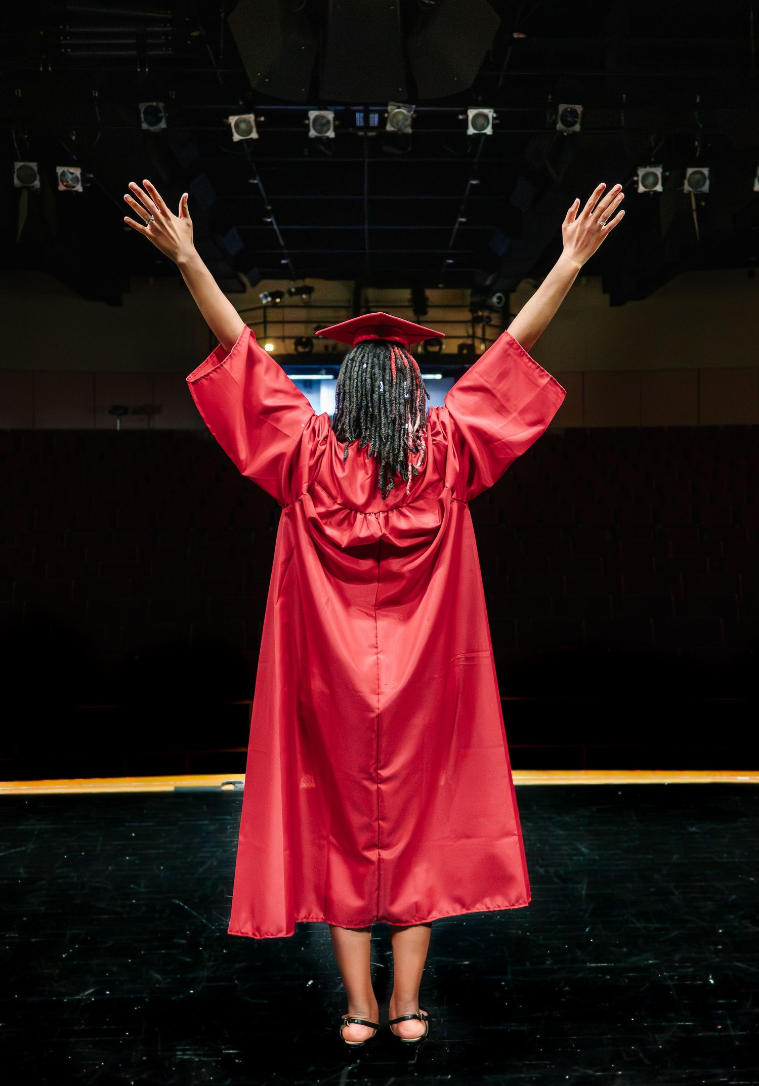 Washington high school graduate standing on theater stage with arms raised in red cap and gown