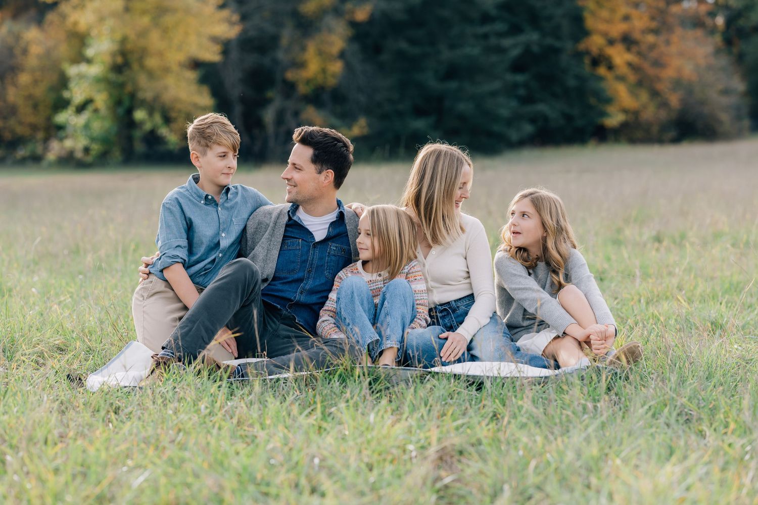 Family of five sitting together on a blanket in a grassy field surrounded by autumn trees.