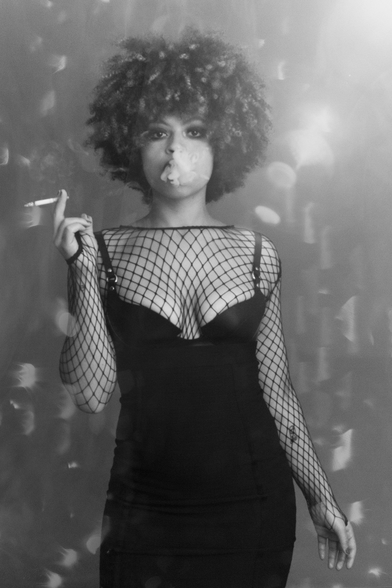 Black and white portrait of woman with afro in fishnet top blowing smoke from cigarette.