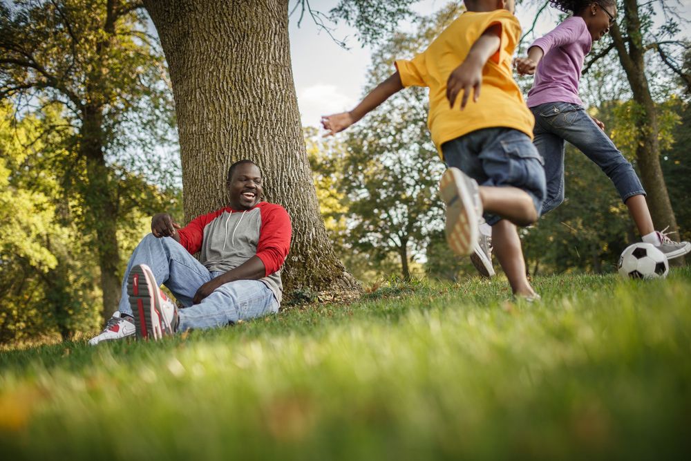 Man relaxing against tree laughing while children play soccer on green grass in sunny park.