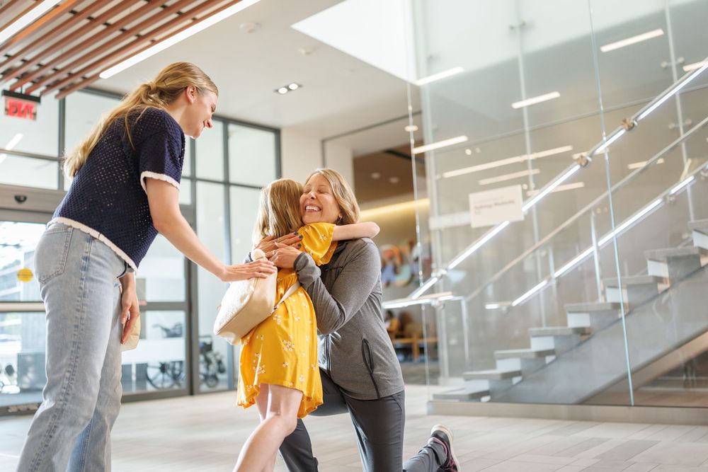 Woman kneeling to hug young girl warmly in bright modern building while another woman watches.