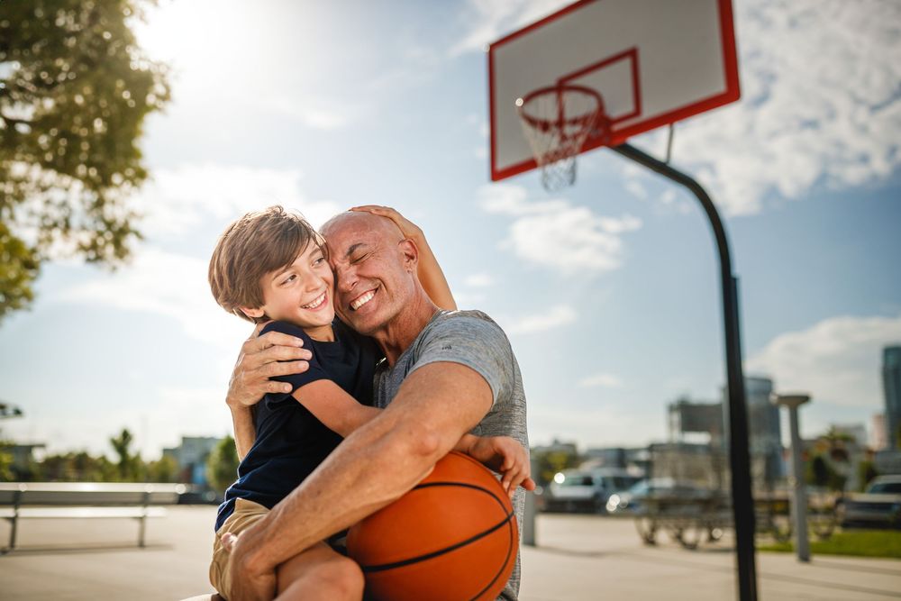 Man and boy share a joyful hug on an outdoor basketball court, holding a basketball with a hoop in the background.
