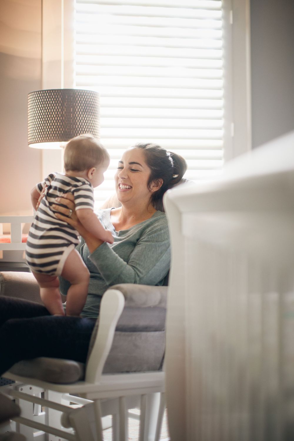 A smiling mother holds her baby in a striped onesie while sitting in a nursery rocking chair by a sunlit window.