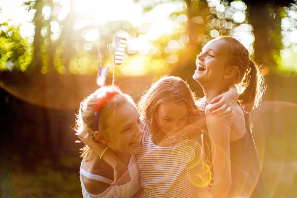 Three joyful girls laughing and playing outdoors in golden sunlight among green trees.