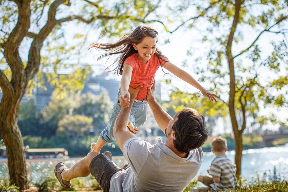 Father joyfully lifting his laughing daughter in a sunny park with trees and a lake in the background.