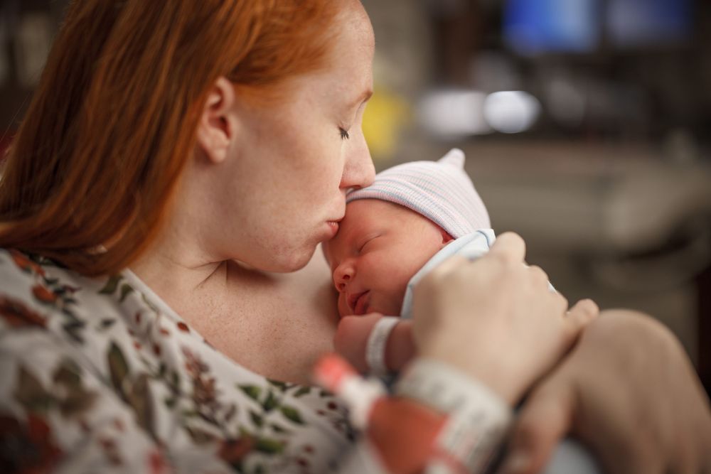 Red-haired mother tenderly kissing her newborn baby in a hospital room.