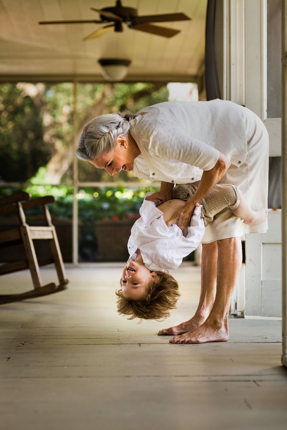 Elderly grandparent playfully holding laughing child upside down on a sunny porch.