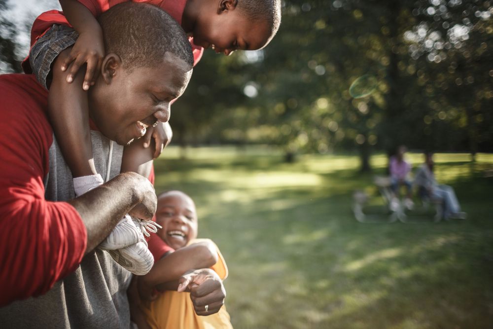 A joyful Black father laughs with two young children in a sunny park, one child on his shoulders.